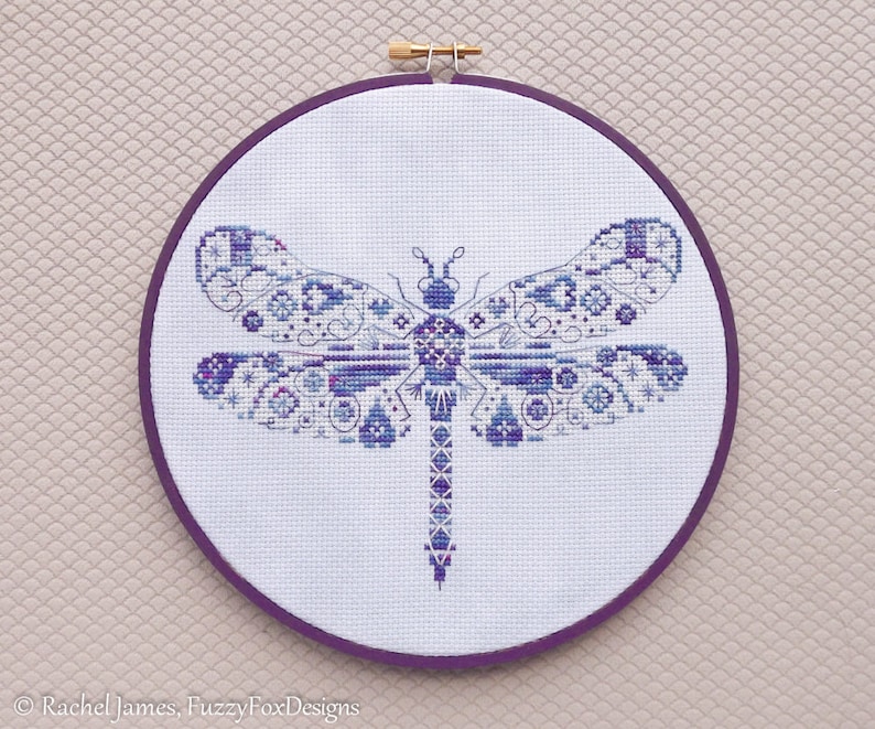 K&ouml;nnte beinhalten: Eine gestickte Libelle in Lila und Wei&szlig; mit Kreuzstich in einem runden Holzrahmen mit einem violetten Stoffr&uuml;cken.