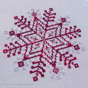 May include: A cross-stitch pattern of a snowflake in shades of pink and purple on white Aida cloth.