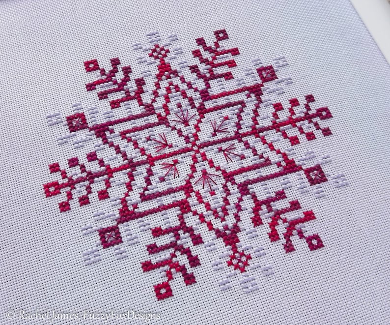 May include: A cross-stitch pattern of a snowflake in shades of pink and purple on white Aida cloth.