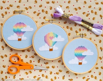 Könnte beinhalten: Drei Stickrahmen mit Kreuzstich-Motiven von Regenbogen-Heißluftballons mit Wolken. Die Rahmen liegen auf einem braunen Stoffhintergrund mit weißem und violettem Stickgarn.