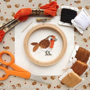 May include: A cross-stitch kit featuring a robin design. The kit includes an embroidery hoop, scissors, needles, and spools of black, white, and brown thread. Orange embroidery floss is also present. The robin is stitched on white fabric.