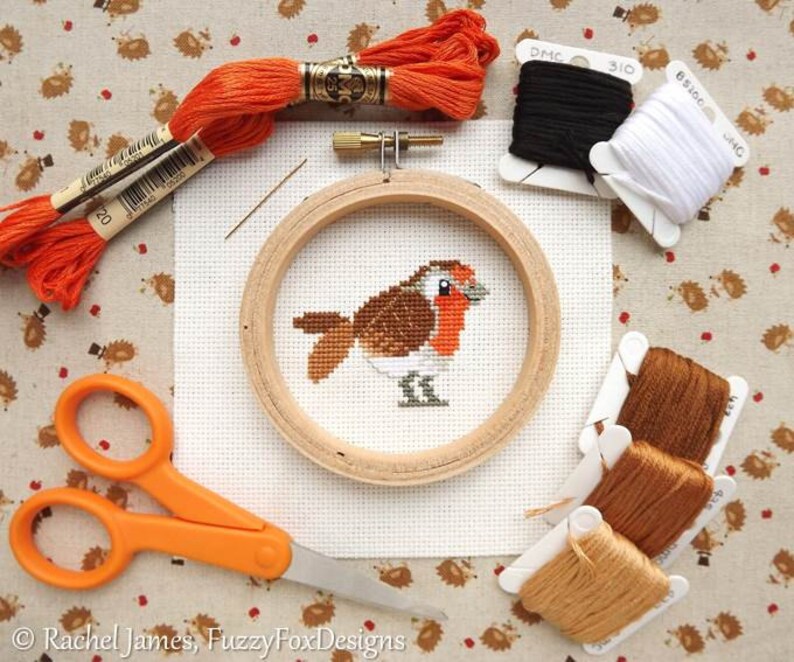 May include: A cross-stitch kit featuring a robin design. The kit includes an embroidery hoop, scissors, needles, and spools of black, white, and brown thread. Orange embroidery floss is also present. The robin is stitched on white fabric.