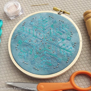 May include: A blue and green cross stitch design of a snowflake with silver beads, framed in a wooden embroidery hoop. The hoop is on a grey textured surface with a spool of thread, a small jar of beads, a pair of scissors, and a wooden needle minder.