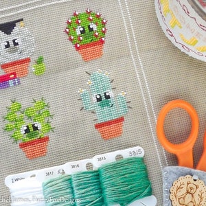 Könnte beinhalten: Kreuzstichmuster mit vier Cartoon-Kakteen in Terrakottatöpfen. Die Kakteen sind grün und blau mit lächelnden Gesichtern. Das Bild enthält auch grünes Stickgarn, eine Schere und ein Filzanhänger mit einem Bärenmotiv.