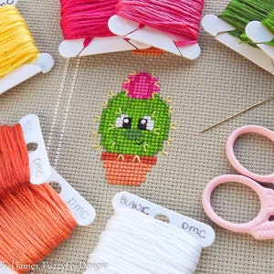 Könnte beinhalten: Ein Kreuzstichmuster eines grünen Kaktus mit einer rosa Blüte oben, in einem Topf. Der Kaktus hat ein lächelndes Gesicht und große Augen. Das Bild enthält DMC-Stickgarn in verschiedenen Farben, eine Nadel und eine rosa Stickerschere.