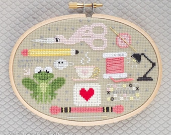 May include: A cross-stitch embroidery hoop with a light gray background featuring a variety of colorful designs, including a frog, a flamingo, a cup of tea, a spool of thread, a pencil, a needle, and a lamp. The text "Happy Little Stitches" is included in the design.