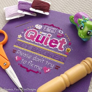 May include: Purple cross-stitch embroidery with the text "Quiet, I'm trying to fix me. Please don't try to fix me."  The embroidery is surrounded by a yellow border and includes a lightbulb, a cup of coffee, and hearts.