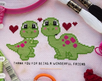 May include: Cross stitch pattern featuring two green dinosaurs with pink hearts, surrounded by pink hearts. The text "THANK YOU FOR BEING A WONDERFUL FRIEND" is stitched below the dinosaurs.