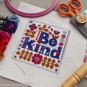 May include: A cross-stitch embroidery project with the phrase "Be Kind" surrounded by colorful flowers and hearts. The project is on white Aida cloth and is framed in a pink embroidery hoop. The image also includes embroidery floss, a needle, and a wooden embroidery hoop.