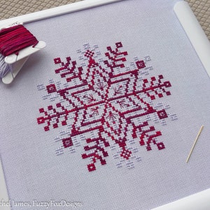 May include: A close-up of a finished cross-stitch project depicting a snowflake in shades of pink and white. The snowflake is stitched on white Aida cloth and framed in a white wooden frame. The project is complete with a needle and floss.