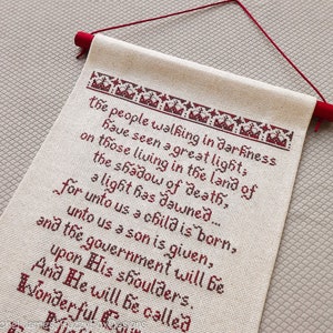 May include: A white cross-stitch sampler with red embroidery depicting a Christmas verse. The verse reads: "The people walking in darkness have seen a great light, on those living in the land of the shadow of death, for unto us a child is born, a light has dawned.... unto us a son is given, and the government will be upon His shoulders. And He will be called Wonderful..."