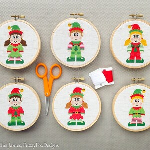 May include: Six cross-stitch elf decorations in wooden hoops, each with a unique elf design in red and green outfits. A pair of orange scissors and a spool of white and red thread are also present.