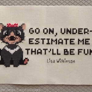 Può includere: Ricamo a punto croce con un diavolo della Tasmania di cartone animato con un fiocco rosso, seduto e sorridente. Il testo recita "Go on, under-estimate me. That'll be fun!" con la firma "Lisa Wilkinson" in basso.