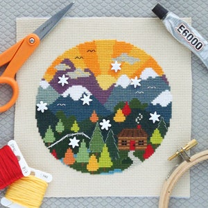 K&ouml;nnte beinhalten: Ein fertiggestelltes Kreuzstich-Stickprojekt, das eine farbenfrohe Bergszene mit einer H&uuml;tte, B&auml;umen und Sternen darstellt. Die Stickerei ist in einem h&ouml;lzernen Stickrahmen montiert. Das Bild zeigt auch Stickgarn, eine Schere und Kleber.