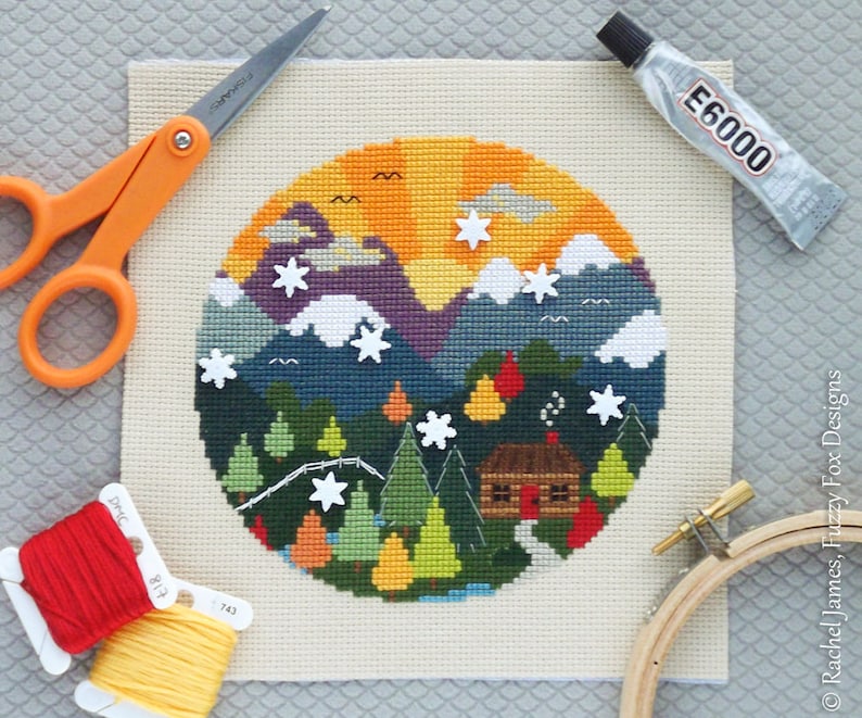 K&ouml;nnte beinhalten: Ein fertiggestelltes Kreuzstich-Stickprojekt, das eine farbenfrohe Bergszene mit einer H&uuml;tte, B&auml;umen und Sternen darstellt. Die Stickerei ist in einem h&ouml;lzernen Stickrahmen montiert. Das Bild zeigt auch Stickgarn, eine Schere und Kleber.