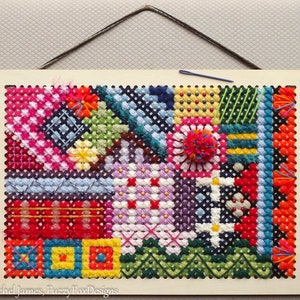 May include: A colorful cross-stitch piece with a patchwork design. The piece is framed in a white wood frame and has a hanging wire.