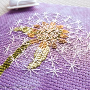 May include: A close-up of a purple cross-stitch embroidery of a dandelion with a green stem and white seeds.