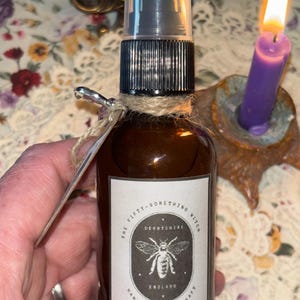 May include: A brown glass bottle with a black spray top and a white label. The label has a black and white illustration of a bee with the text "The Fifty-Something Witch Derbyshire England Handmade Spellcraft".