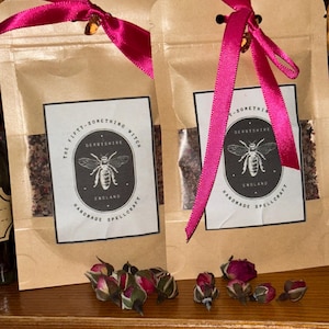May include: Two small brown paper bags tied with pink ribbon. Each bag contains a small amount of loose brown and white material. The bags have black and white labels with the text "The Fifty-Something Witch", "Derbyshire", "England", and "Handmade Spellcraft".