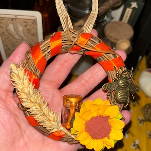 May include: A small circular wreath made of brown twigs wrapped with orange ribbon. The wreath is decorated with a stalk of dried wheat, a yellow sunflower, a small glass jar with gold-coloured honey, and a metallic bronze-toned bee charm.