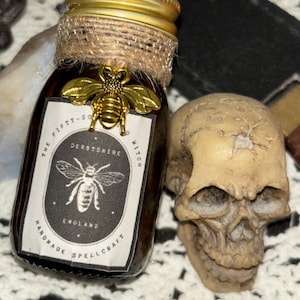 May include: A small glass bottle with a gold lid and a brown label. The label says "The Fifty-Something Derbyshire Witch England Handmade Spellcraft". There is a gold bee charm on the bottle.