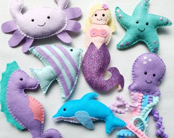 May include: A set of felt sea creature toys, including a purple crab, a turquoise and purple fish, a purple and pink mermaid, a turquoise starfish, a purple octopus with pink tentacles, a purple seahorse, and a blue dolphin.