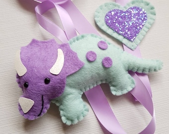 May include: A purple and green felt triceratops with a white horn and black eyes. A purple glittery heart is attached to a lavender ribbon.