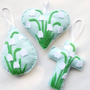 May include: Three felt ornaments in the shape of a heart, a teardrop, and a cross. Each ornament is light blue with white flowers and green stems.