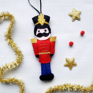 May include: A felt Christmas ornament depicting a toy soldier. The soldier is wearing a red and gold uniform with a blue hat and black hair. The ornament is hanging from a string.