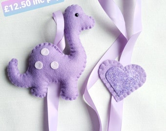 May include: A lavender felt dinosaur ornament with white polka dots and a matching heart-shaped ornament. Both are attached to lavender satin ribbons. The dinosaur has a long neck and a small black eye.