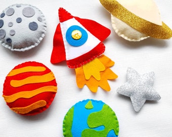 May include: A set of felt toys featuring a rocket, planets, a star, and a ringed planet. The rocket is red, white, and orange. The planets are orange, red, blue, and gray. The star is silver. The ringed planet is cream with gold glitter.
