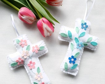 May include: Two white felt cross ornaments with embroidered floral designs. One cross has pink flowers and leaves, the other has blue flowers and leaves.