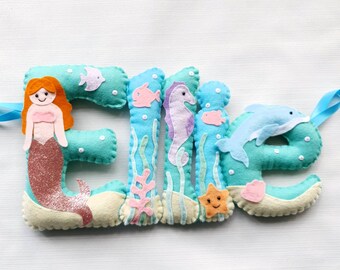 May include: A felt name banner with the name "Ellie" spelled out in turquoise letters. Each letter is decorated with a sea-themed design, including a mermaid, a seahorse, a starfish, and a dolphin.