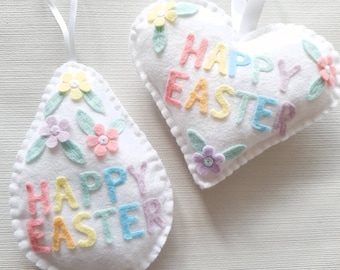 May include: Two white felt ornaments, one shaped like an egg and the other like a heart, both decorated with pastel colored felt flowers and leaves. The ornaments say "Happy Easter" in pastel colored felt letters.