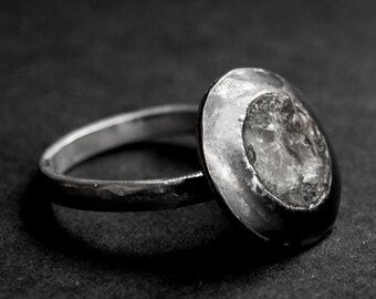 May include: A silver ring with a round, textured, silver setting. The setting has a slightly raised, circular, white stone.