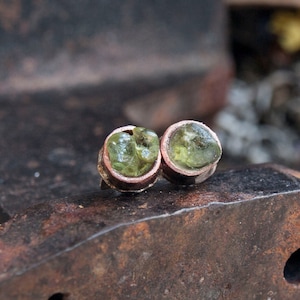 May include: A pair of stud earrings with raw, light green peridot gemstones set in copper. The earrings are displayed on a weathered metal surface, highlighting their natural beauty. The earrings are a unique jewelry item. The gemstones are a vibrant green.