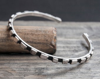 May include: A silver bracelet with a square-shaped design. The bracelet is on a wooden surface.