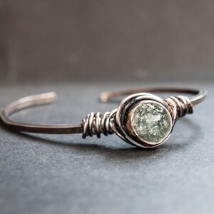 Puede incluir: Una pulsera de alambre plateado con una piedra preciosa verde. La piedra preciosa est&aacute; engastada en un engaste de plata y est&aacute; rodeada de alambre de plata.