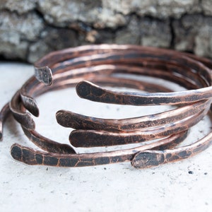 Può includere: Cinque braccialetti in rame martellato impilati uno sull'altro. I braccialetti sono di un colore rame caldo con una finitura rustica e martellata.