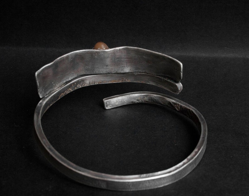 K&ouml;nnte beinhalten: Ein silbernes Armband mit einem einzigartigen, geschwungenen Design. Das Armband hat eine glatte, polierte Oberfl&auml;che und einen einzigen kleinen braunen Stein als Akzent.
