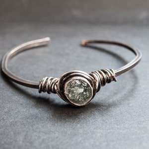 Puede incluir: Un brazalete de alambre plateado con una piedra redonda verde clara engastada en el centro.