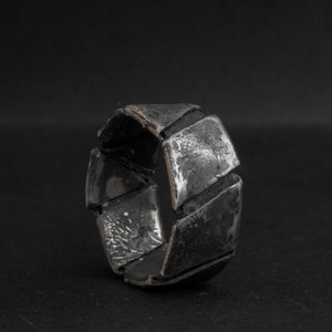 Könnte beinhalten: Nahaufnahme eines silbernen Rings mit geometrischem Design. Der Ring besteht aus mehreren rechteckigen Segmenten, die einen strukturierten Look erzeugen. Das Metall hat eine dunkle, oxidierte Oberfläche, die dem Schmuck Tiefe verleiht.