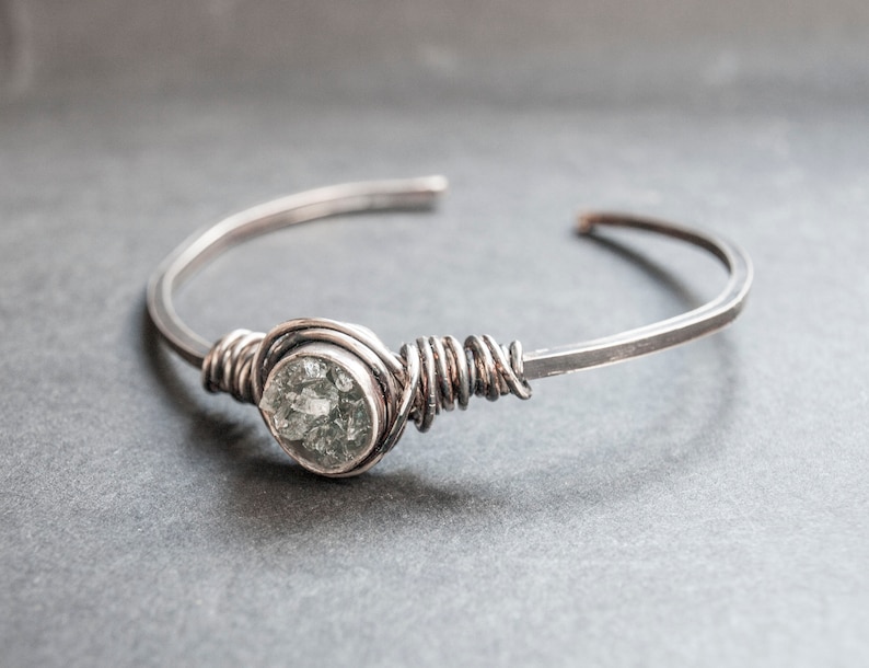 Puede incluir: Un brazalete de alambre plateado con una piedra preciosa transparente y facetada.