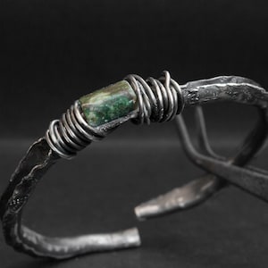 May include: A handcrafted silver cuff bracelet with a textured finish. The bracelet features a rectangular green stone wrapped in silver wire. The open-ended design allows for easy wear. The bracelet's width is approximately 1.3 cm.
