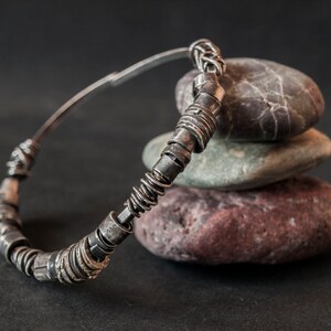 May include: A silver bangle bracelet with a unique design of twisted metal rings. The bracelet is resting on a stack of smooth, gray and brown stones.