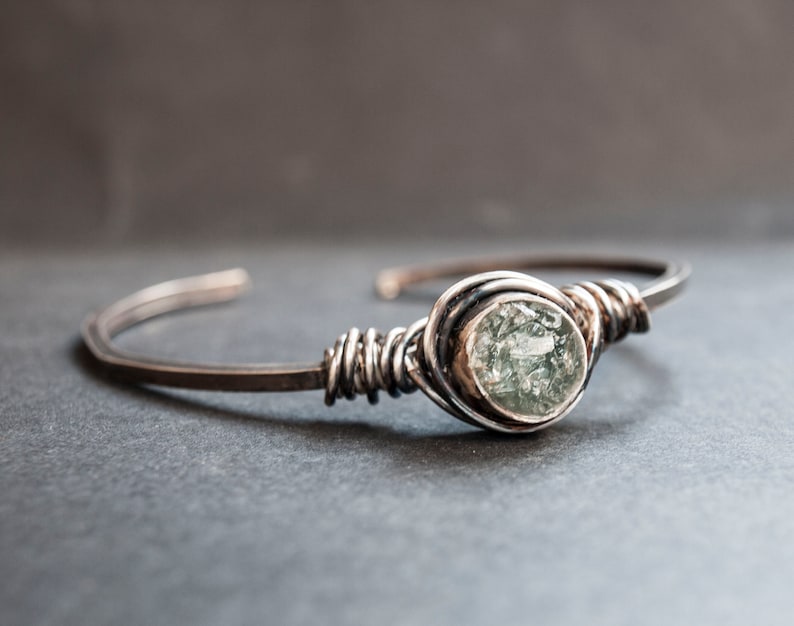 Puede incluir: Una pulsera de alambre plateado con una piedra preciosa verde. La piedra preciosa est&aacute; engastada en un engaste de plata y est&aacute; rodeada de alambre de plata.