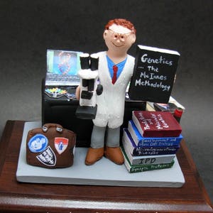 May include: A figurine of a scientist holding a microscope and a chalkboard with the text "Genetics... The Me Innes Methodology". The scientist is wearing a white lab coat and a red tie. There are books stacked on a shelf behind the scientist, including books titled "Paedy tries", "Molecular Genetics", "Development and Stem Cell Biology", "Neurodegenerative Diseases", "IPD", and "Synaptic Proteins".