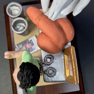 K&ouml;nnte beinhalten: Eine Keramikfigur einer Person mit braunen Haaren, die ein gr&uuml;nes Hemd tr&auml;gt und ein Paar Handschellen h&auml;lt. Die Figur steht auf einer wei&szlig;en Oberfl&auml;che mit einem braunen, orangefarbenen und wei&szlig;en Keramikobjekt, einem kleinen Bilderrahmen und zwei grauen Beh&auml;ltern.