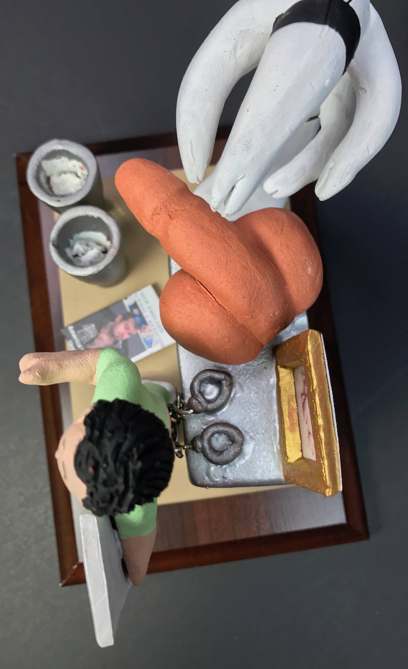 K&ouml;nnte beinhalten: Eine Keramikfigur einer Person mit braunen Haaren, die ein gr&uuml;nes Hemd tr&auml;gt und ein Paar Handschellen h&auml;lt. Die Figur steht auf einer wei&szlig;en Oberfl&auml;che mit einem braunen, orangefarbenen und wei&szlig;en Keramikobjekt, einem kleinen Bilderrahmen und zwei grauen Beh&auml;ltern.