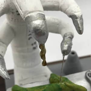 K&ouml;nnte beinhalten: Ein wei&szlig;er Roboterarm mit einer silbernen Spitze h&auml;lt einen Bohrer &uuml;ber einem gr&uuml;nen, strukturierten Objekt. Der Roboterarm ist &uuml;ber einem Tisch mit einer silbernen Oberfl&auml;che positioniert.